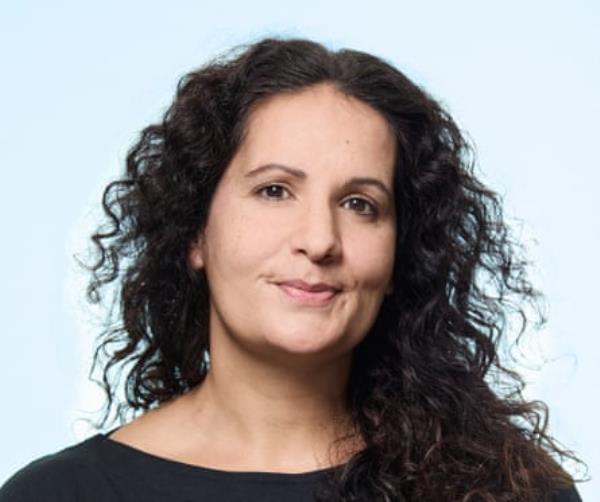 A middle-aged woamn with dark wavy hair, wearing a black, round-neck top, shot against a pale blue background