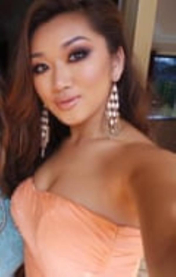 A young woman with long dark hair, wearing full makeup, large, dangly earrings and a peach strapless dress