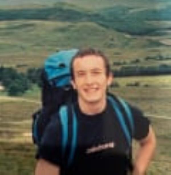 A young man with short brown hair, wearing a sports top and carrying a rucksack, walking on a hillside