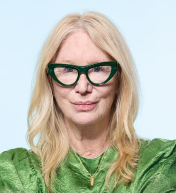 An older woman with blo<em></em>nde hair and large-f<em></em>ramed, green cat-eye glasses, wearing a shiny green dress and a gold necklace, shot against a pale blue background