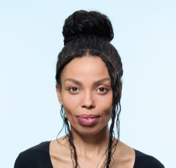 A portrait of Emma Dabiri with her hair in a bun on top of her head and some loose small plaits, wearing several necklaces and a black V-neck T-shirt, shot against a pale blue background