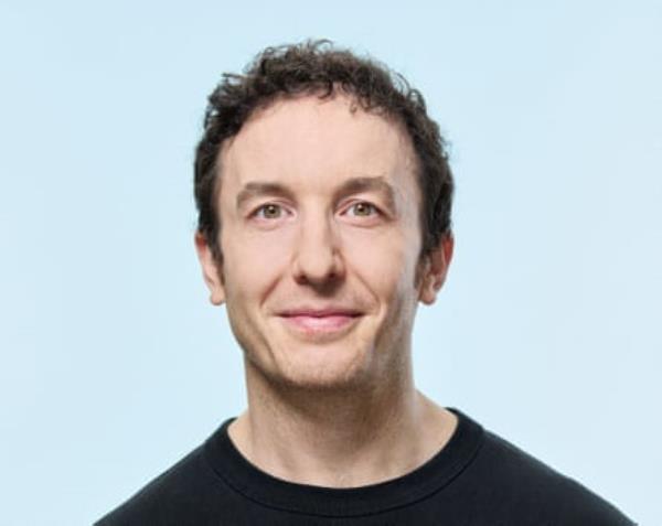 A portrait of a middle-aged man with brown hair, wearing a black T-shirt, shot against a pale blue background