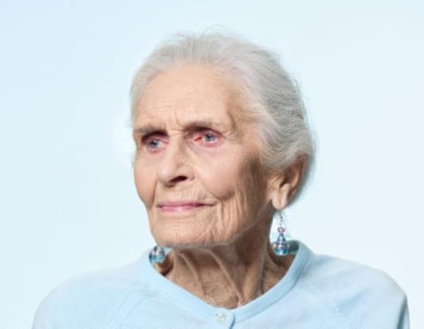 A portrait of an elderly lady with white hair pulled back from her face, dangly earrings and a pale blue cardigan, shot against a pale blue background