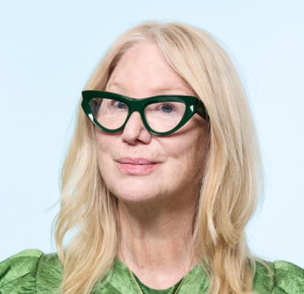 An older woman with blo<em></em>nde hair and large-f<em></em>ramed, green cat-eye glasses, wearing a shiny green dress and a gold necklace, shot against a pale blue background