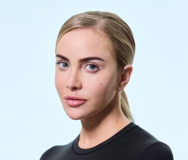 A young blo<em></em>nde woman with her hair tied back, wearing a black, round-neck top, shot against a pale blue background