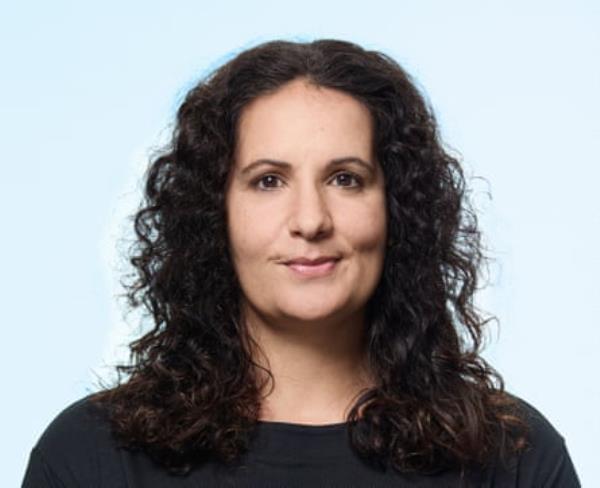 A middle-aged woamn with dark wavy hair, wearing a black, round-neck top, shot against a pale blue background